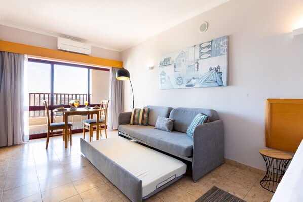 A compact living space with a sofa, a pull-out bed, a dining area, and a balcony.
