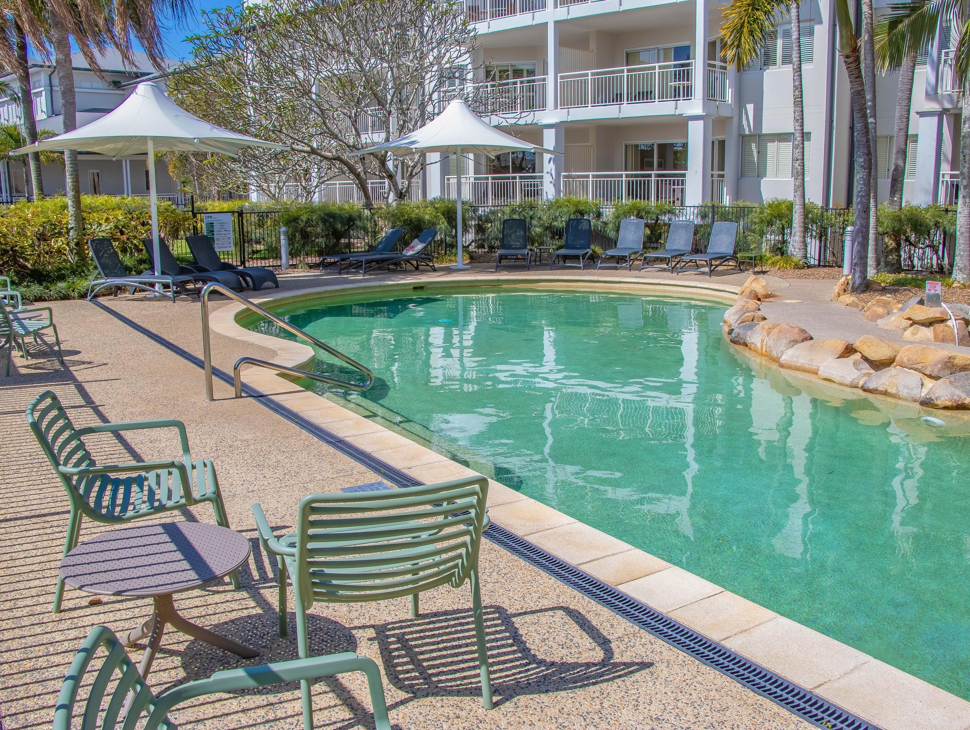 Piscina com espreguiçadeiras e guarda-sóis, cercada por área ajardinada com palmeiras.