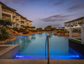A swimming pool with a rock-lined edge, surrounded by a resort building with balconies and outdoor seating areas. - The Pool Resort Kingscliff - 1 BR Ocean view (2320) - Holiday Management Co. (Tweed)
