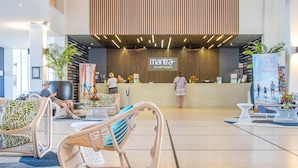 Zona de recepción de hotel con diseño moderno, que incluye un mostrador de recepción, área de estar con sillas de mimbre y un cartel grande que anuncia 'Mantra on Salt Beach'.