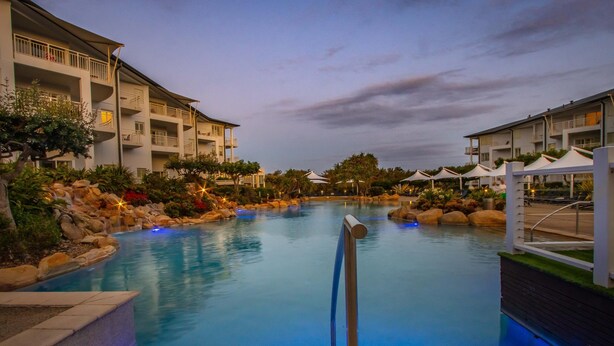 A swimming pool with a rock-lined edge, surrounded by a resort building with balconies and outdoor seating areas. - The Pool Resort Kingscliff - 2B Ocean View (2320/21) - Holiday Management Co. (Tweed)
