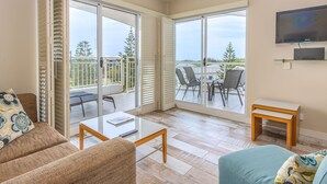 A living room with a glass table, two sofas, a TV, and a balcony with chairs and a table. - The Pool Resort Kingscliff - 2B Ocean View (2320/21) - Holiday Management Co. (Tweed)