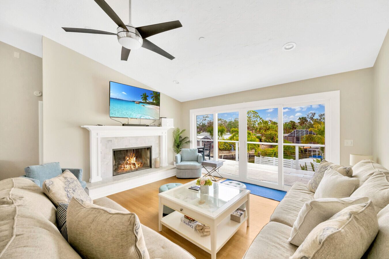 A spacious living room with a fireplace, a ceiling fan, and a large sliding glass door leading to an outdoor deck with a view of a tropical beach.
