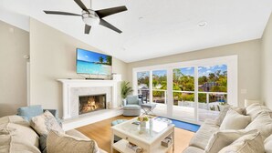Ein großzügiges Wohnzimmer mit Kamin, Deckenventilator und einer großen Schiebetür zu einer Terrasse mit Blick auf einen tropischen Strand.