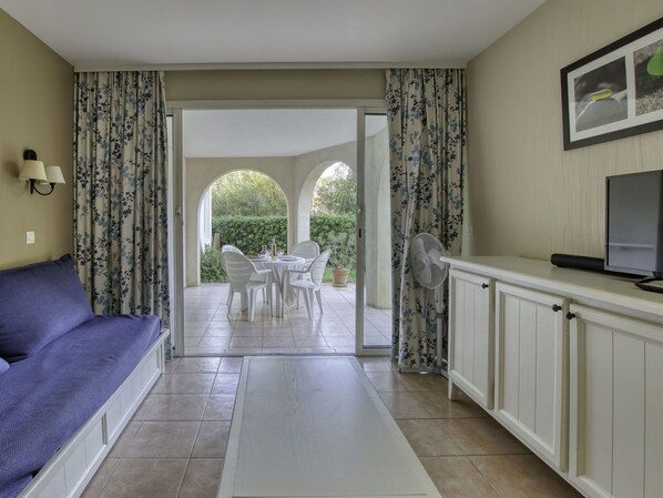 A room with a sofa, a dining table with chairs, a TV, and a view of a garden through an open doorway.