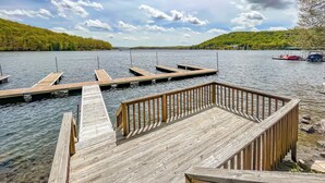 A wooden dock extending into a calm lake with clear water, surrounded by lush green hills and a partly cloudy sky.