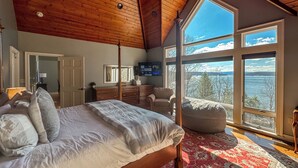A bedroom with a large bed, a wooden ceiling, a red patterned rug, and a view of a lake through large windows.