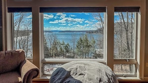 A room with a large window overlooking a lake and trees, a brown chair, and a white pillow.