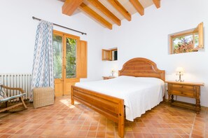 A bedroom with a wooden bed, a nightstand, a chair, a small table, and a window with a view of trees. - YourHouse Son Jaumell 6, villa with pool in Cala Agulla (Capdepera)