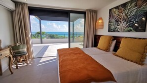 Ein Schlafzimmer mit einem großen Bett, einem Schreibtisch und einem Stuhl. Durch die Glastüren hat man Blick aufs Meer.