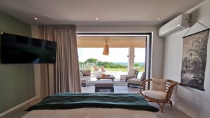 Ein Schlafzimmer mit Bett, Wandfernseher, großem Fenster mit Vorhängen, einem Sessel und Blick ins Freie.