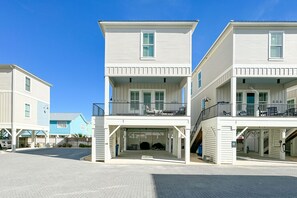 A row of two-story beachfront houses with balconies, a covered parking area, and a clear blue sky. - Steps from beach! Sleeps 10! Bikes, Xbox, BBQ! (Gulf Shores)