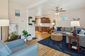 Una sala de estar moderna con un sofá gris, un pouf redondo y una mesa de centro. Se observa una cocina al fondo con armarios de madera y un ventilador de techo.
