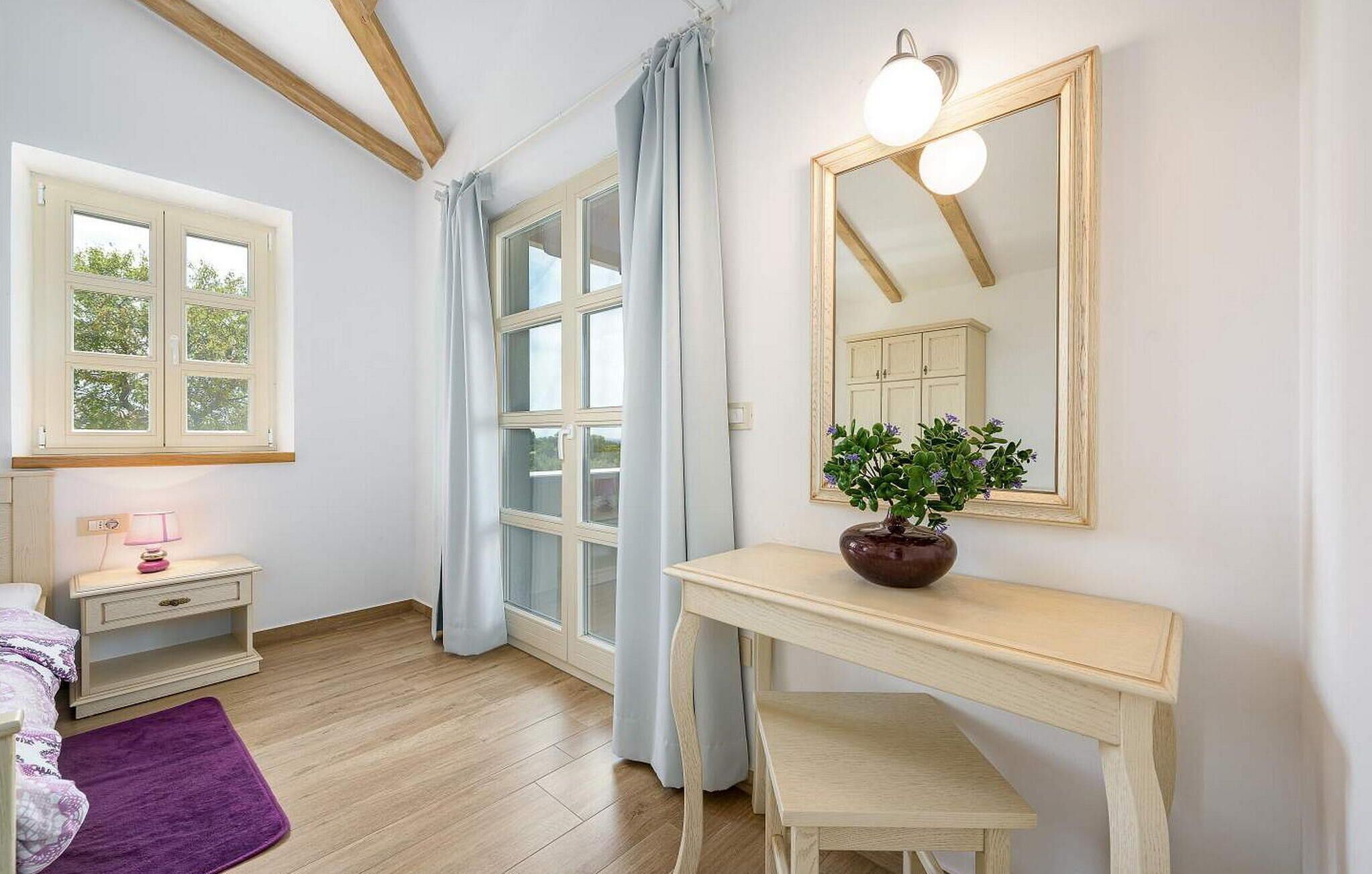 Ein Schlafzimmer mit einem Bett, einem Nachttisch mit Lampe, einer Kommode, einem Spiegel und einem Fenster mit Vorhängen.