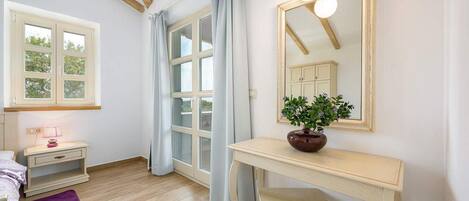 Ein Schlafzimmer mit einem Bett, einem Nachttisch mit Lampe, einer Kommode, einem Spiegel und einem Fenster mit Vorhängen.