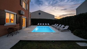Une piscine rectangulaire, agrémentée de chaises longues et d’une table, et entourée d’une haie et de bâtiments.