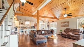 A spacious living area with a wooden ceiling, a large sofa, a recliner, and a dining area with a kitchen in the background.