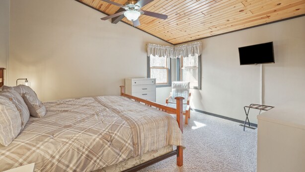 A bedroom with a wooden ceiling, a bed with patterned bedding, a nightstand, a dresser, a chair, a window with curtains, and a TV mounted on the wall.