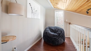 A room with a wooden ceiling, a black bean bag chair, a white railing, and a window with a view of trees.