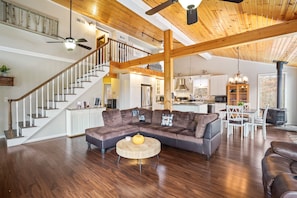 A spacious living area with a wooden ceiling, a large sectional sofa, a round coffee table, and a staircase leading to an upper level.