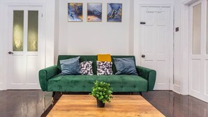 Un sofá verde con cojines estampados, una mesa de centro de madera con una planta, y arte abstracto en la pared.