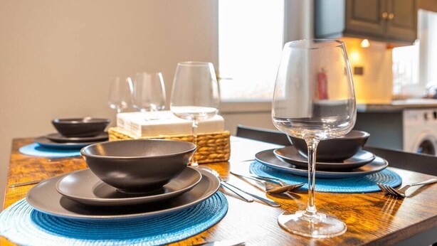 Una mesa de comedor dispuesta con platos, tazones y una copa de vino negros, con manteles individuales azules y una cesta tejida al fondo.