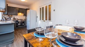 Cocina moderna con armarios oscuros, un refrigerador blanco y un comedor con una mesa de madera dispuesta para dos personas, con copas de vino y fuentes.