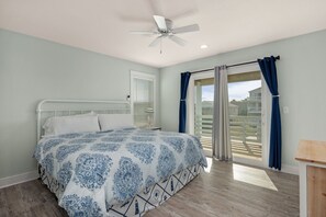 A bedroom with a bed, a ceiling fan, a window with curtains, and a view of a balcony. - Ocean Breeze: Dog-Friendly w/Views, Elevator, Private Pool, Large fenced in Yard (Kitty Hawk)