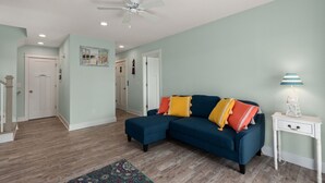 A living room with a blue sectional sofa, a white side table with a lamp, and a patterned rug. - Ocean Breeze: Dog-Friendly w/Views, Elevator, Private Pool, Large fenced in Yard (Kitty Hawk)