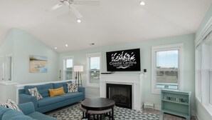 A living room with a blue sofa, a fireplace, and a television displaying 'Coastal Carolina Vacations and Sales'. - Ocean Breeze: Dog-Friendly w/Views, Elevator, Private Pool, Large fenced in Yard (Kitty Hawk)