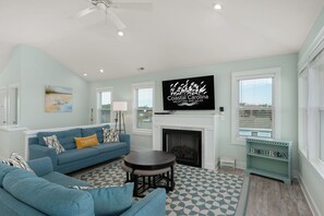 Un salon avec un canapé bleu, une cheminée et un téléviseur diffusant une publicité pour « Coastal Carolina Vacations and Sales ».