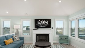 A living room with a fireplace, a blue sofa, a coffee table, and a television displaying 'Coastal Carolina Vacations and Sales'. - Ocean Breeze: Dog-Friendly w/Views, Elevator, Private Pool, Large fenced in Yard (Kitty Hawk)