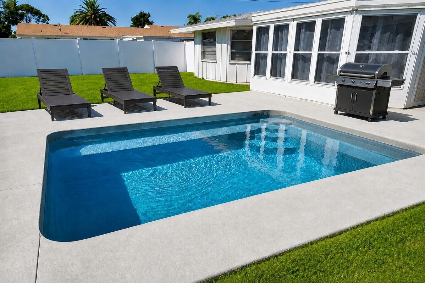 A rectangular swimming pool with a concrete deck, surrounded by a white fence, and equipped with a barbecue grill.