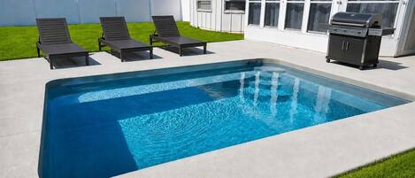 A rectangular swimming pool with a concrete deck, surrounded by a white fence, and equipped with a barbecue grill.