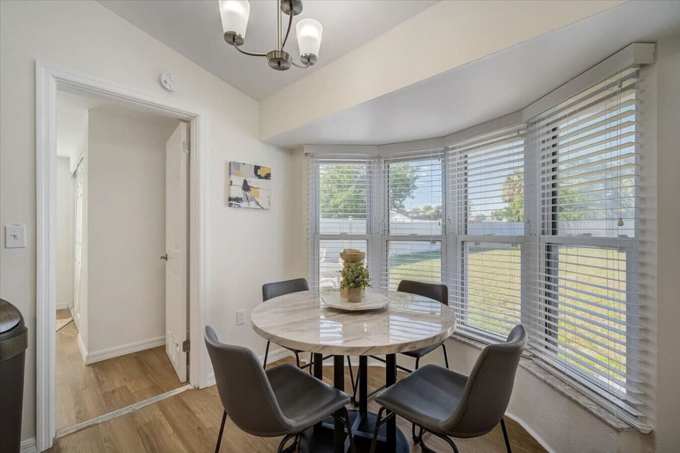 A dining area with a round table, four chairs, a chandelier, and large windows with blinds.