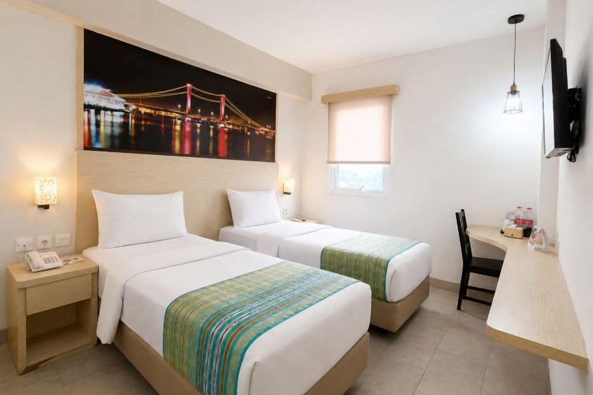 A hotel room with two beds, a desk, a chair, a TV, and a painting of a bridge.