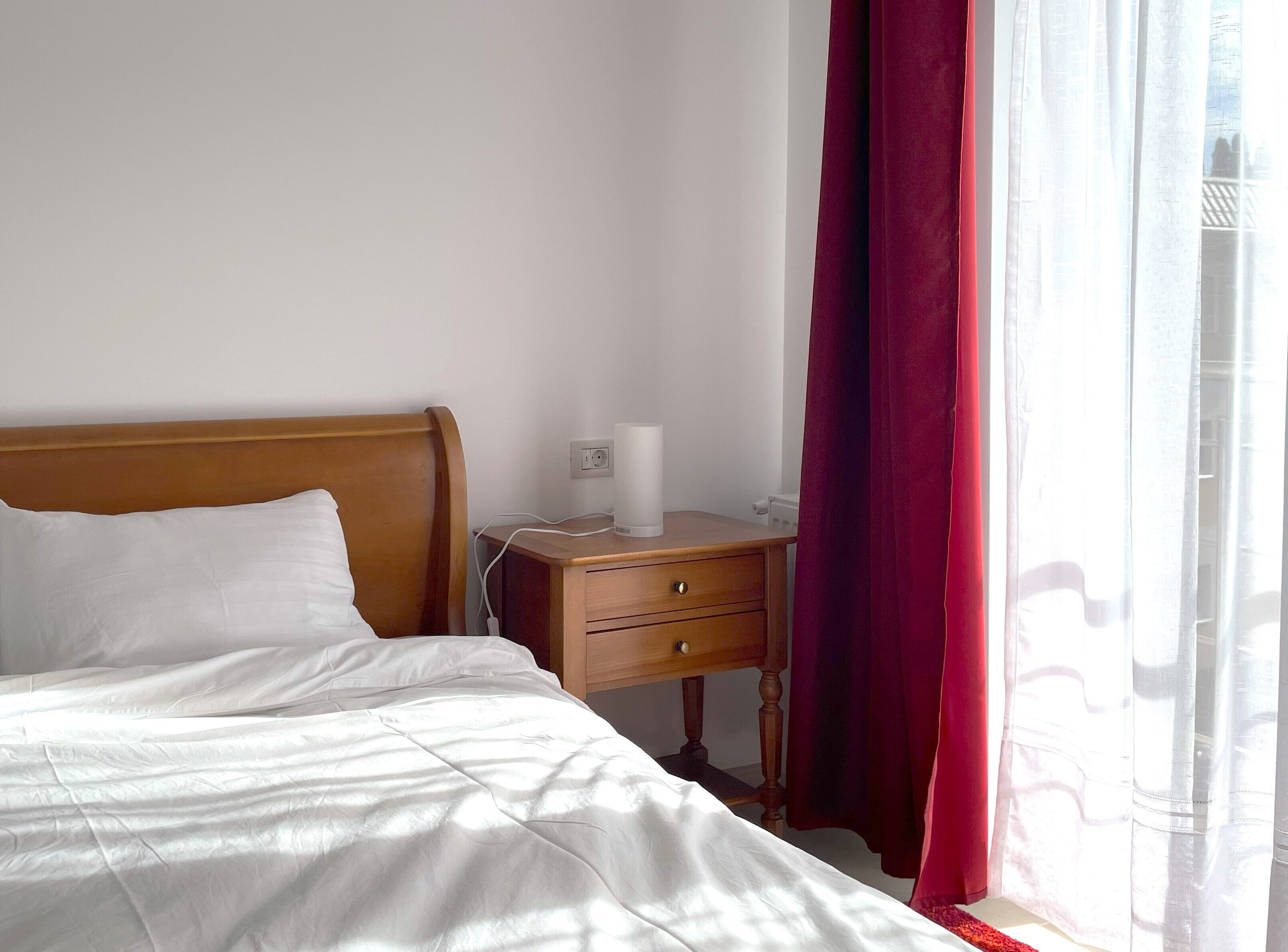 Una cama bien hecha con sábanas blancas, una mesita de noche de madera con una lámpara y una ventana con cortinas translúcidas.