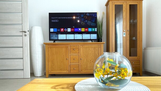 Ein Wohnzimmer mit einem Holzkabinett, einer Glaskaraffe mit gelben Blumen auf einer runden Matte und einem Flachbildfernseher, der einen Homescreen anzeigt.