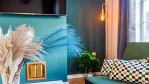 En væg i teal-farve med en dekorativ plante, en vægmonteret lampe og en pudde med et mønster på en grøn sofa.