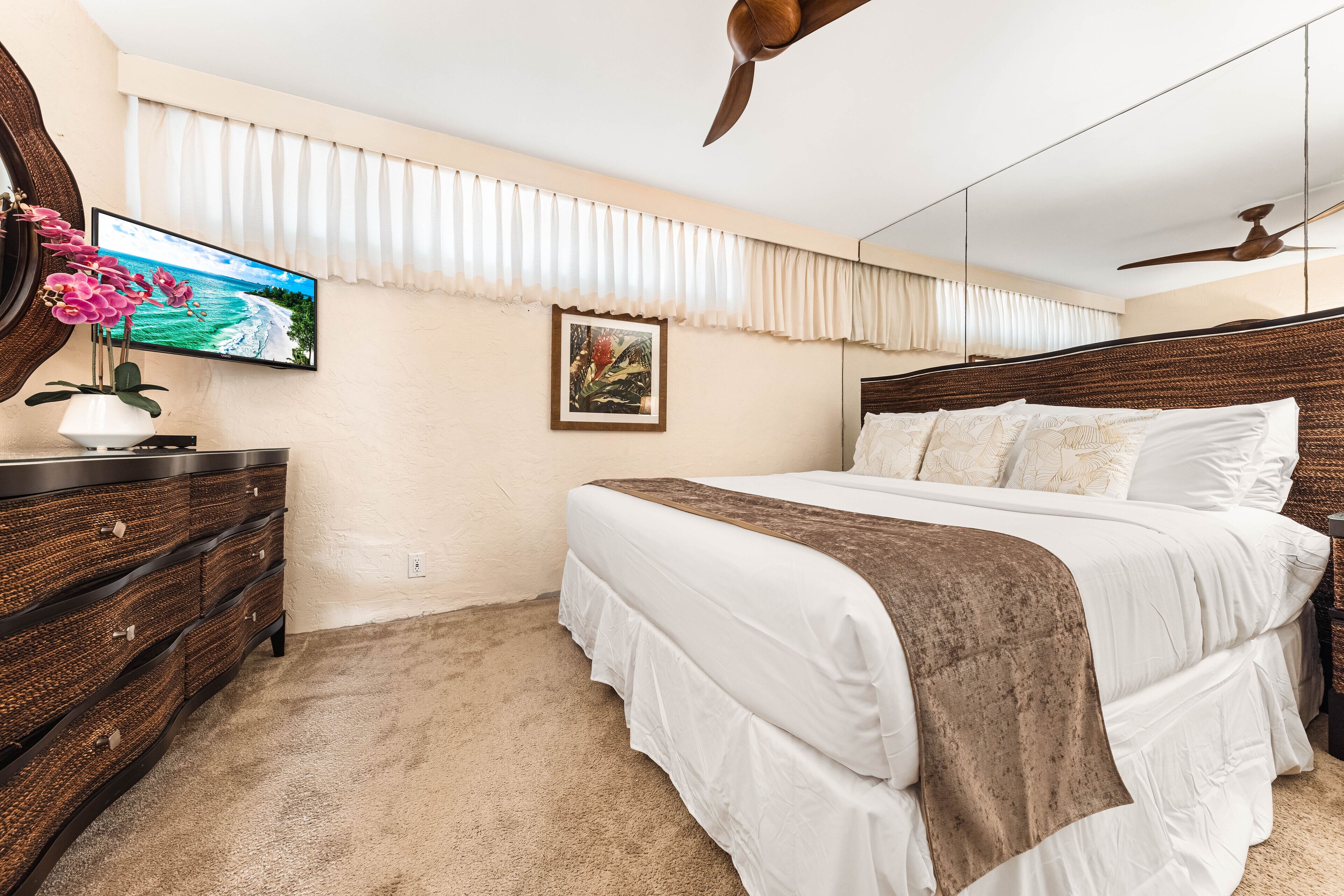 Ein Schlafzimmer mit Bett, Kommode, Fernseher, Deckenventilator und einem Fenster mit Vorhängen.