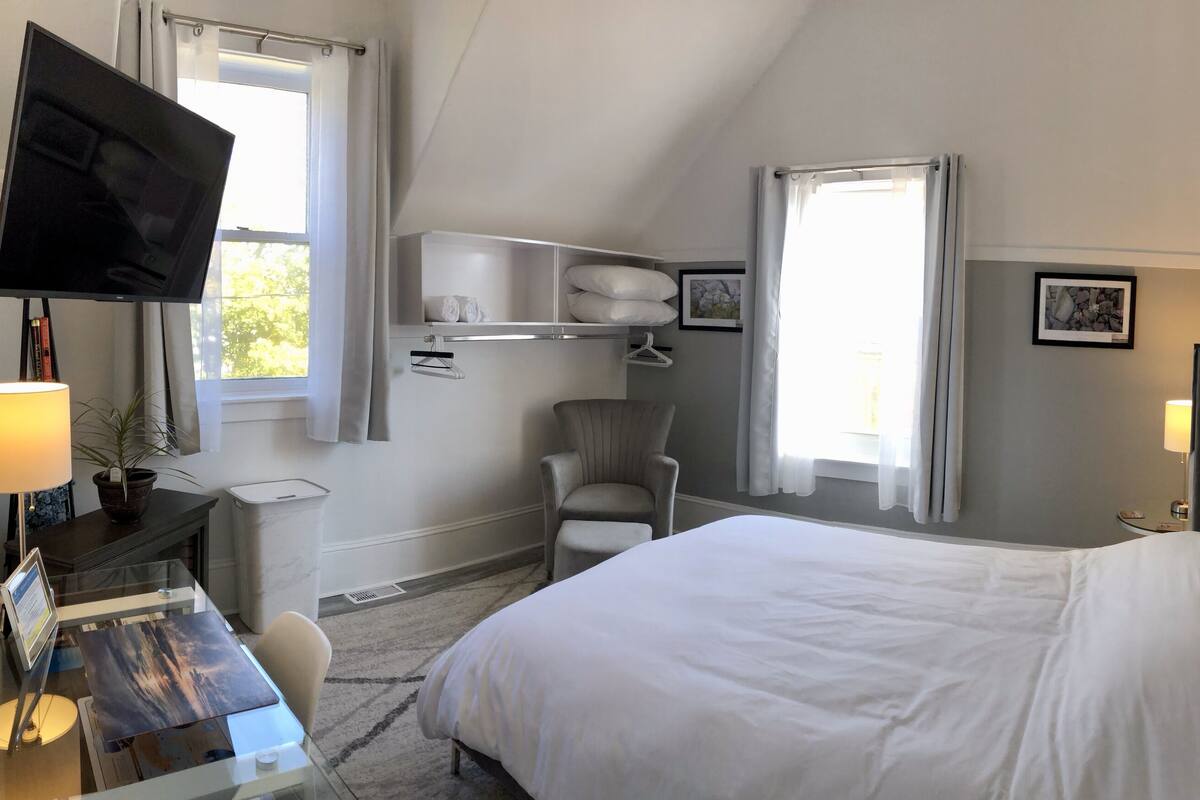 A hotel room with a bed, a desk with a lamp, a chair, a TV mounted on the wall, a shelf with towels, and a painting on the wall.