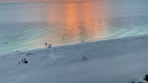 Una playa con gente caminando y de pie, y una puesta de sol sobre el agua.