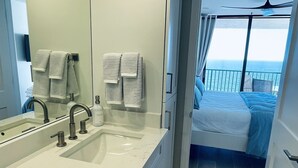 Un baño moderno con un espejo grande, un lavabo blanco y una zona de ducha. Se ve un balcón con una cama y un ventilador de techo.
