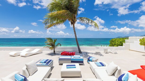 A beachside lounge area with white lounge chairs and blue-striped cushions, a palm tree, and a view of the ocean.
