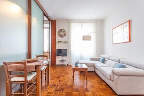 A living room with a wooden floor, a sofa, a dining table with chairs, a TV, and a window with curtains.