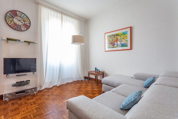A living room with a flat-screen TV, wooden flooring, a sofa set, a lamp, and a wall clock.