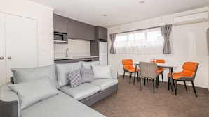 A modern living room with a grey sofa, orange chairs, a dining table, and a kitchen area with grey cabinets and a microwave. - Aurora City Suites Motel (Rotorua)