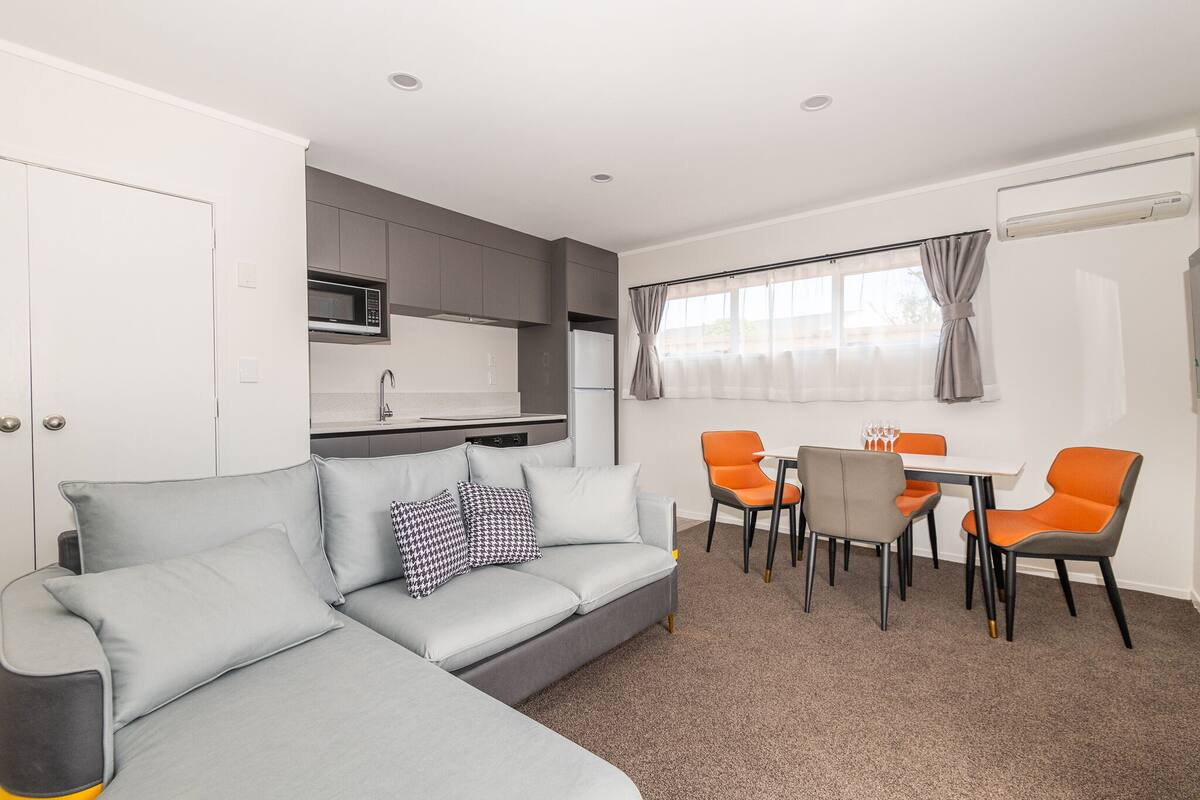 A modern living room with a grey sofa, orange chairs, a dining table, and a kitchen area with grey cabinets and a microwave.