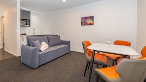 A modern kitchen with a microwave and sink, a gray sofa with cushions, and a dining table with orange chairs. - Aurora City Suites Motel (Rotorua)