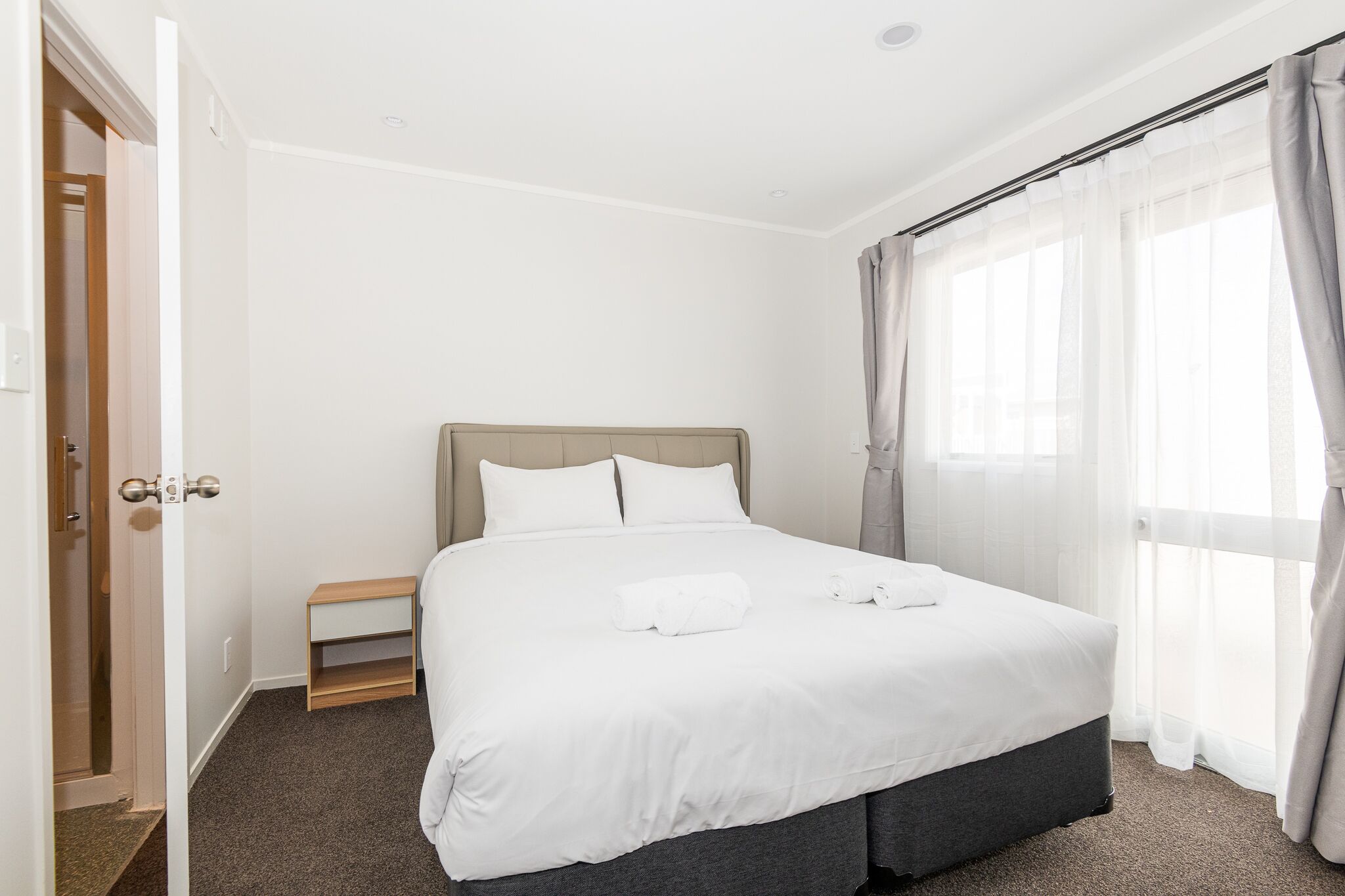 Una cama bien hecha con sábanas blancas, una mesita de noche de madera y una ventana con cortinas traslúcidas.